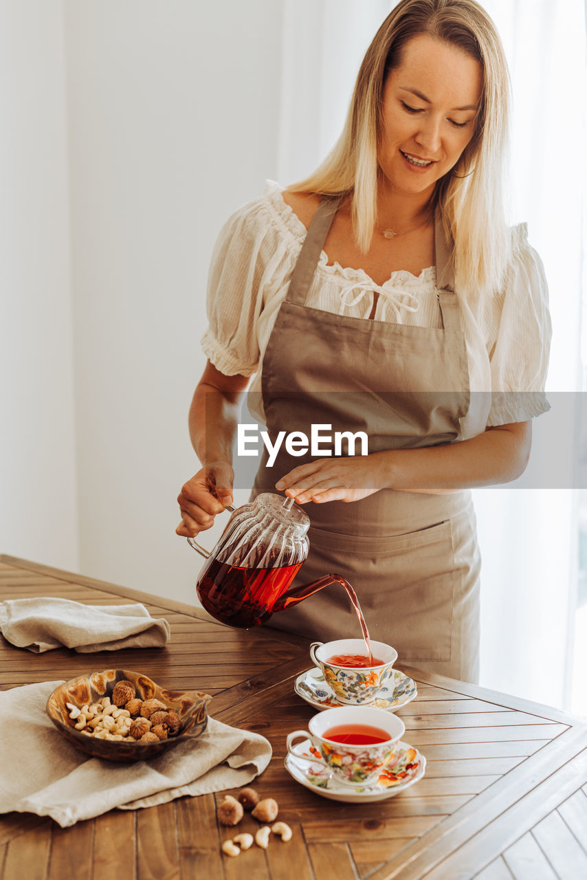 Blond woman in apron pouring hibiscus tea in cups on table