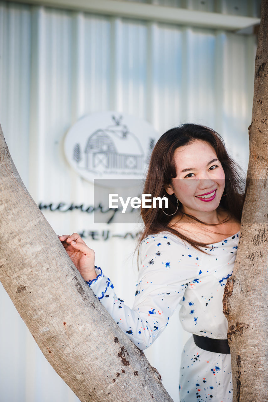 PORTRAIT OF A SMILING YOUNG WOMAN STANDING AGAINST WALL