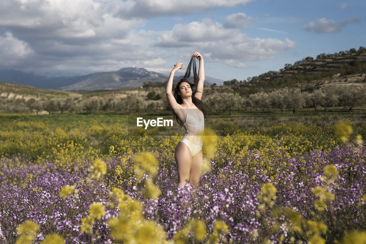 WOMAN STANDING BY FLOWERS ON FIELD AGAINST SKY