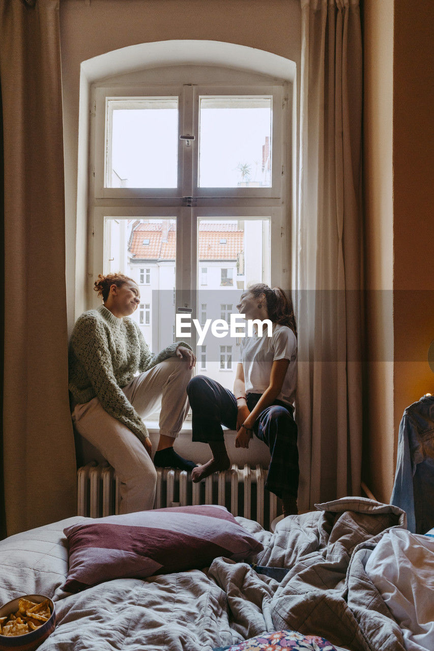 Young female friends talking while sitting at window in bedroom