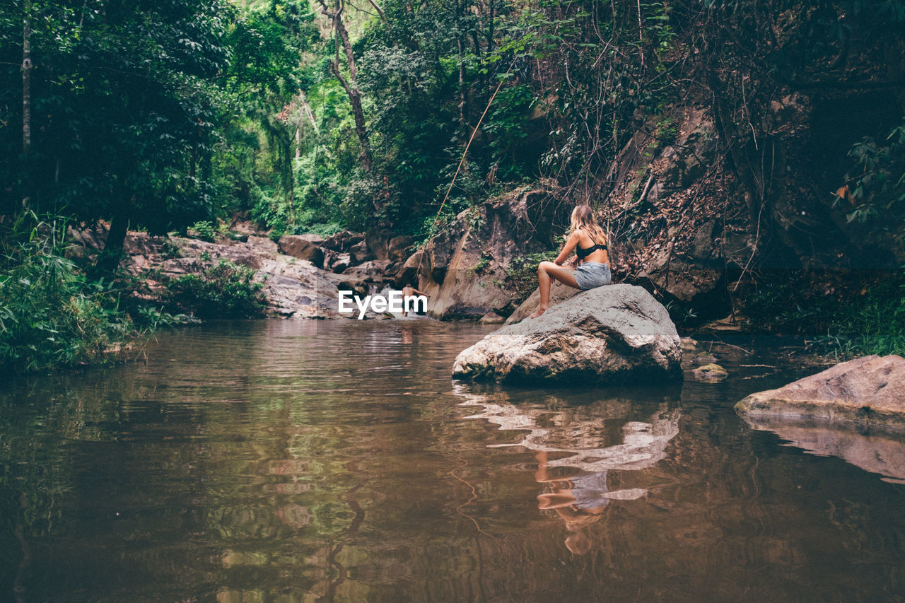 Rear view of woman sitting on rock at stream in forest