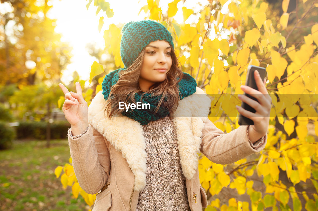 Young woman in warm clothing taking selfie through smart phone while showing peace sign against autumn tree