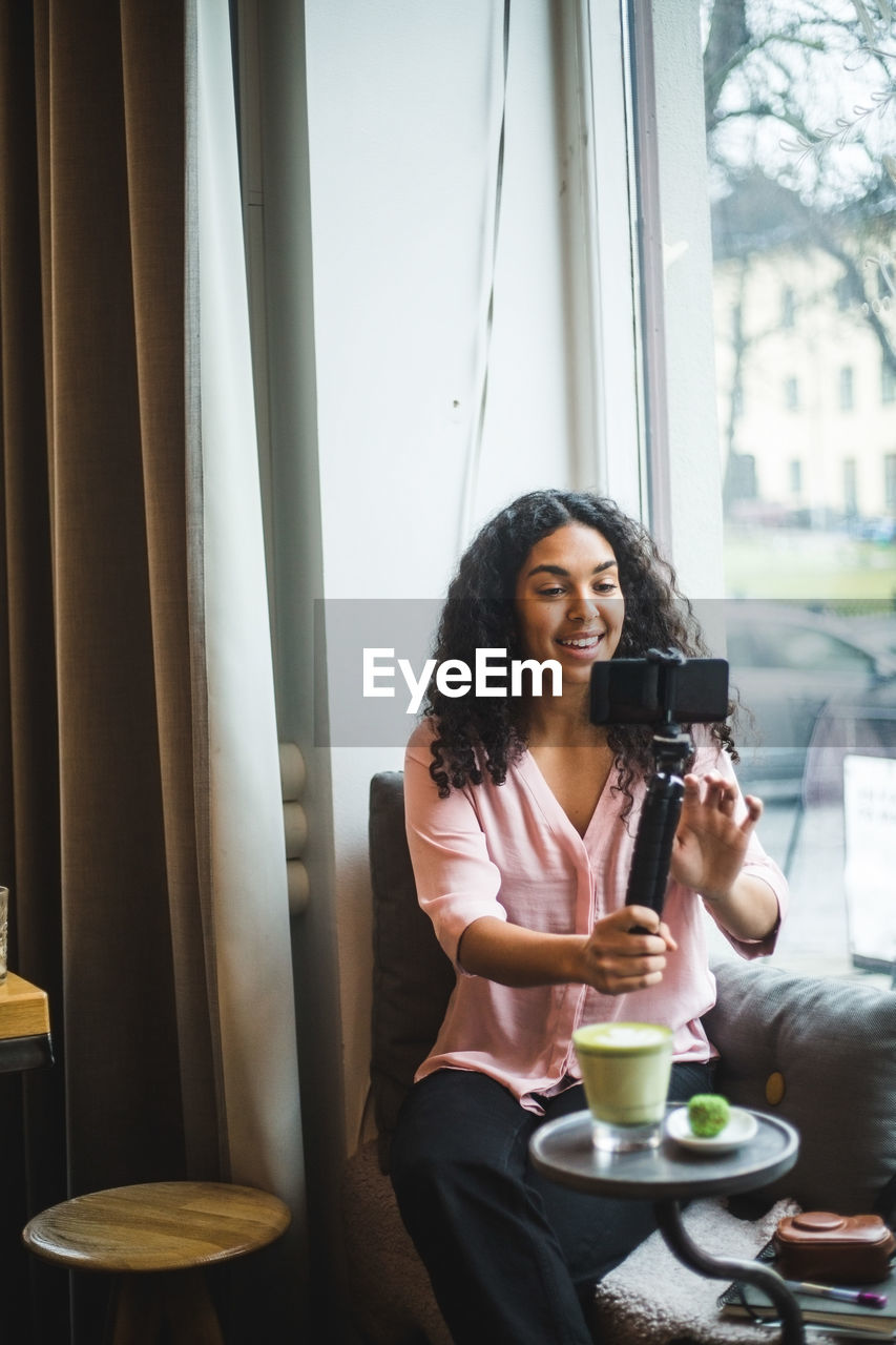 Smiling young female influencer vlogging through mobile phone on tripod sitting by window at creative office