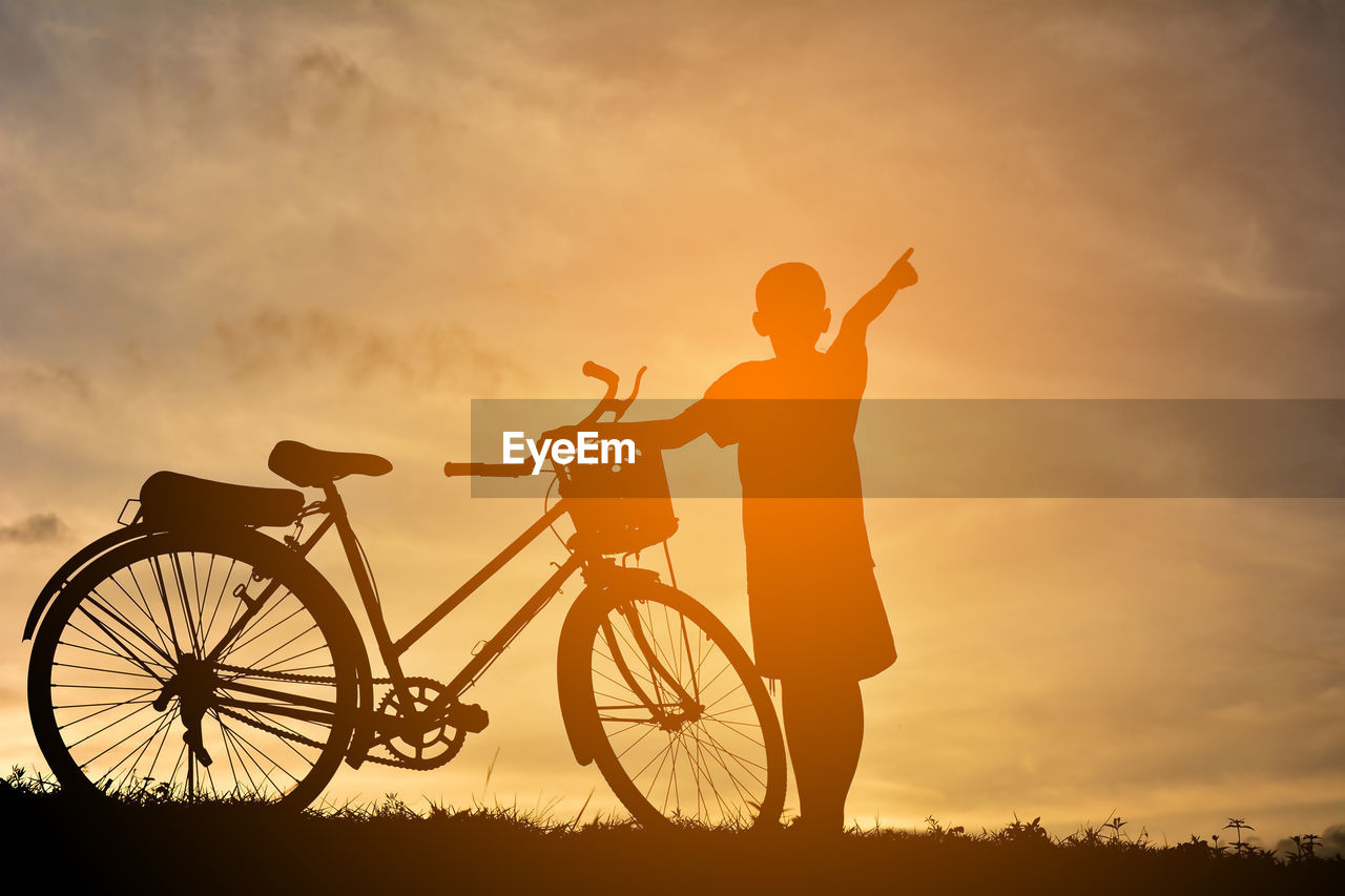 Silhouette boy with bicycle pointing towards sky while standing on field during sunset