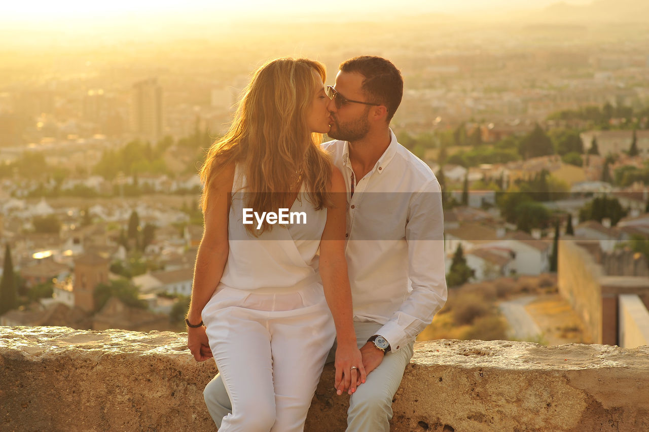 Young romantic couple kissing while sitting on retaining wall against cityscape