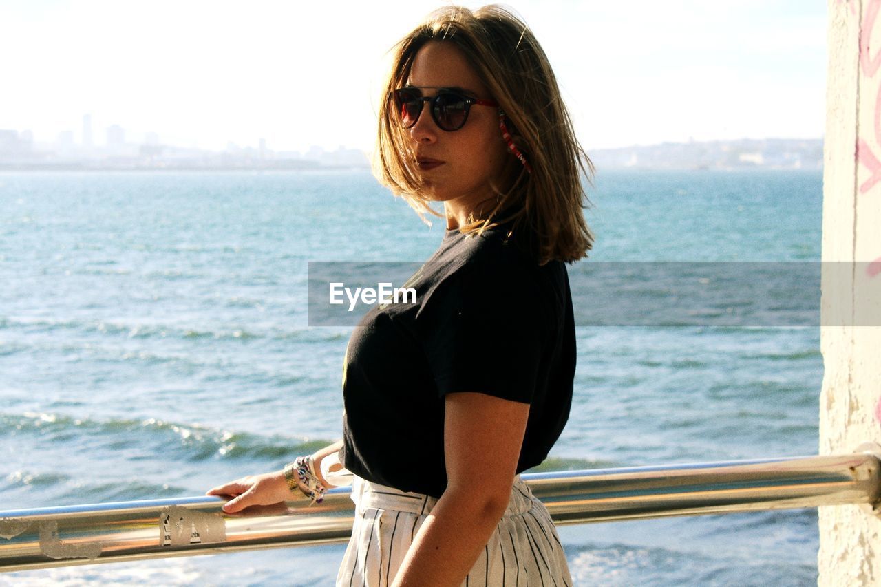 Portrait of young woman wearing sunglasses standing by sea against sky