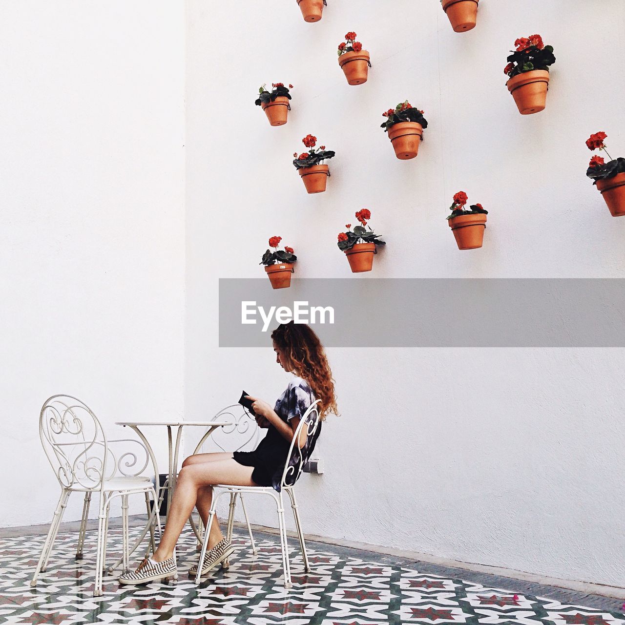 Side view of young woman sitting on chair looking at menu card against potted plants on wall indoors
