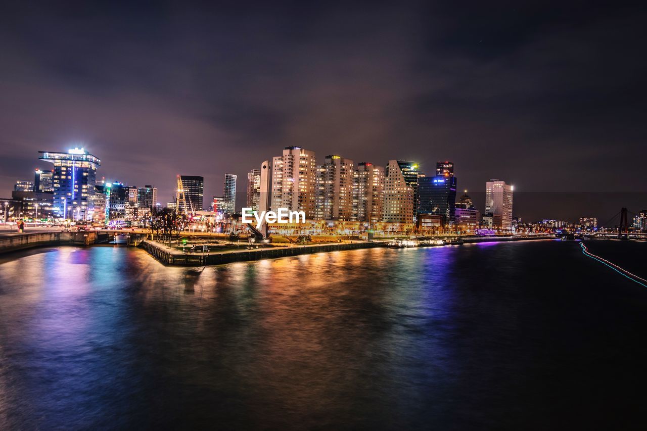 Illuminated buildings by river against sky at night