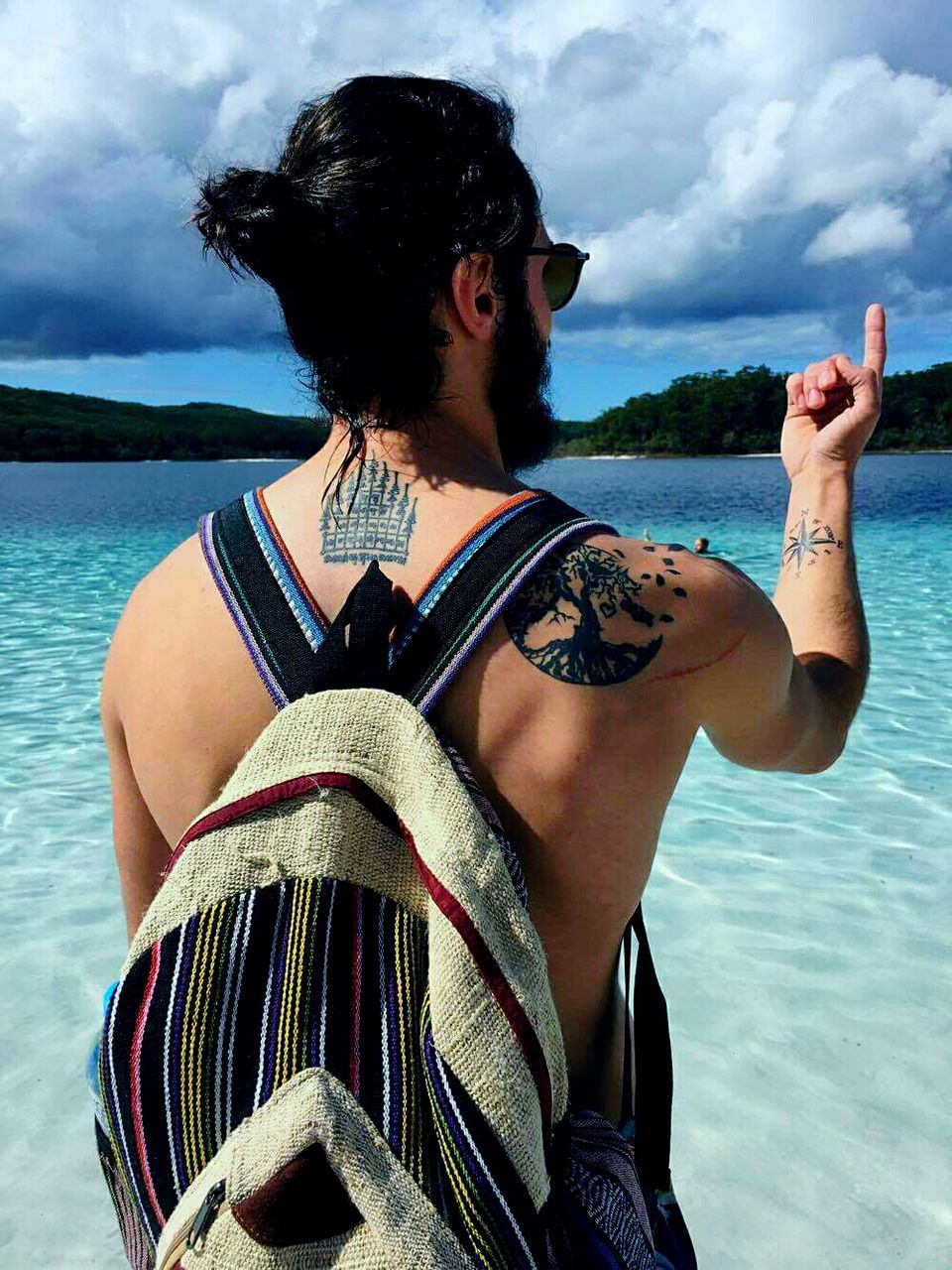 Rear view of shirtless man with backpack pointing at beach against cloudy sky