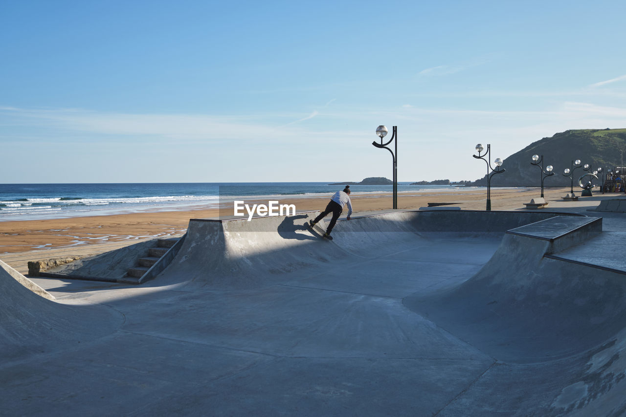 Unrecognizable teen boy riding skateboard in skate park on sunny day on seashore