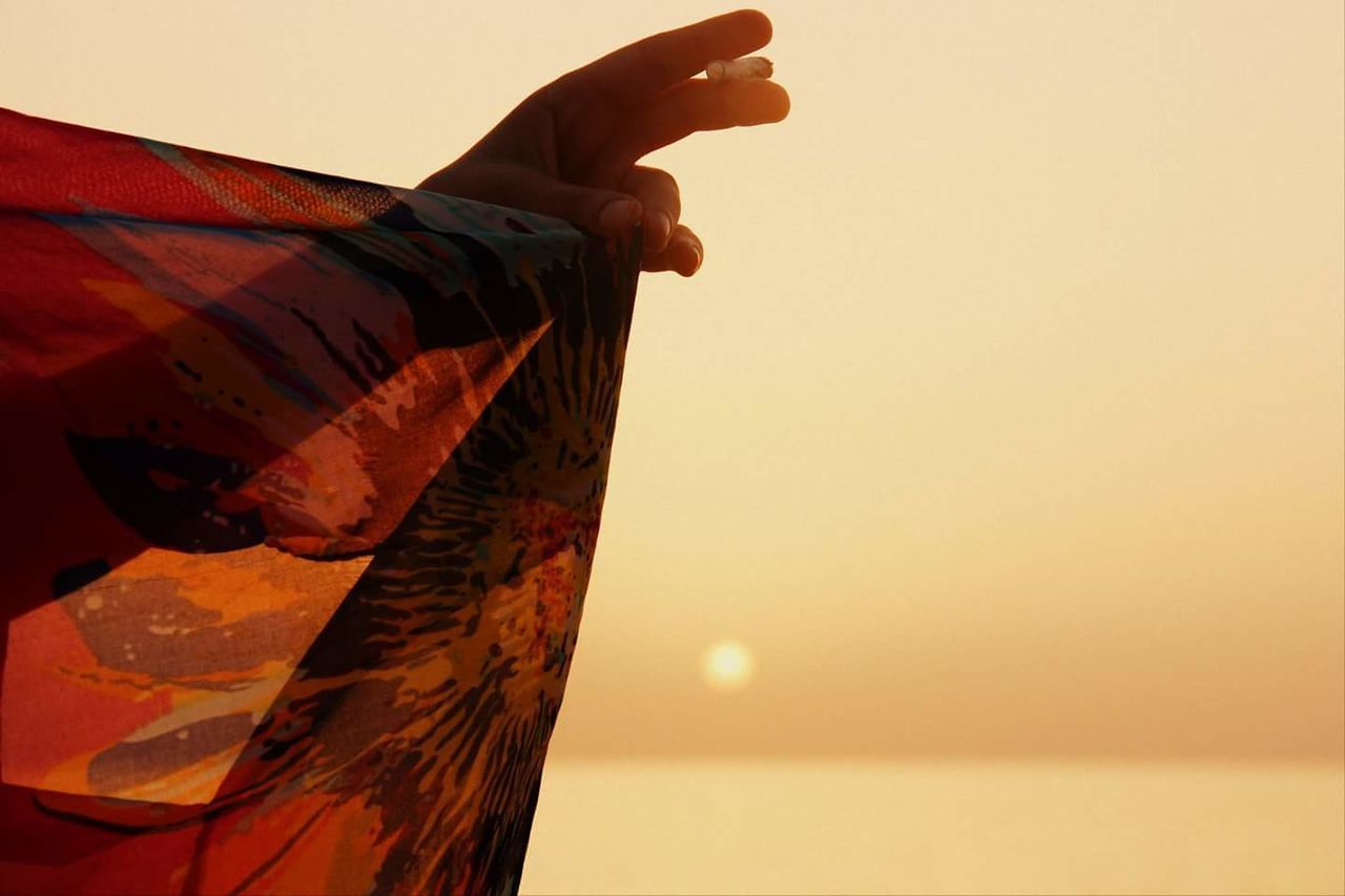 Cropped hand of woman holding scarf and cigarette at beach during sunset