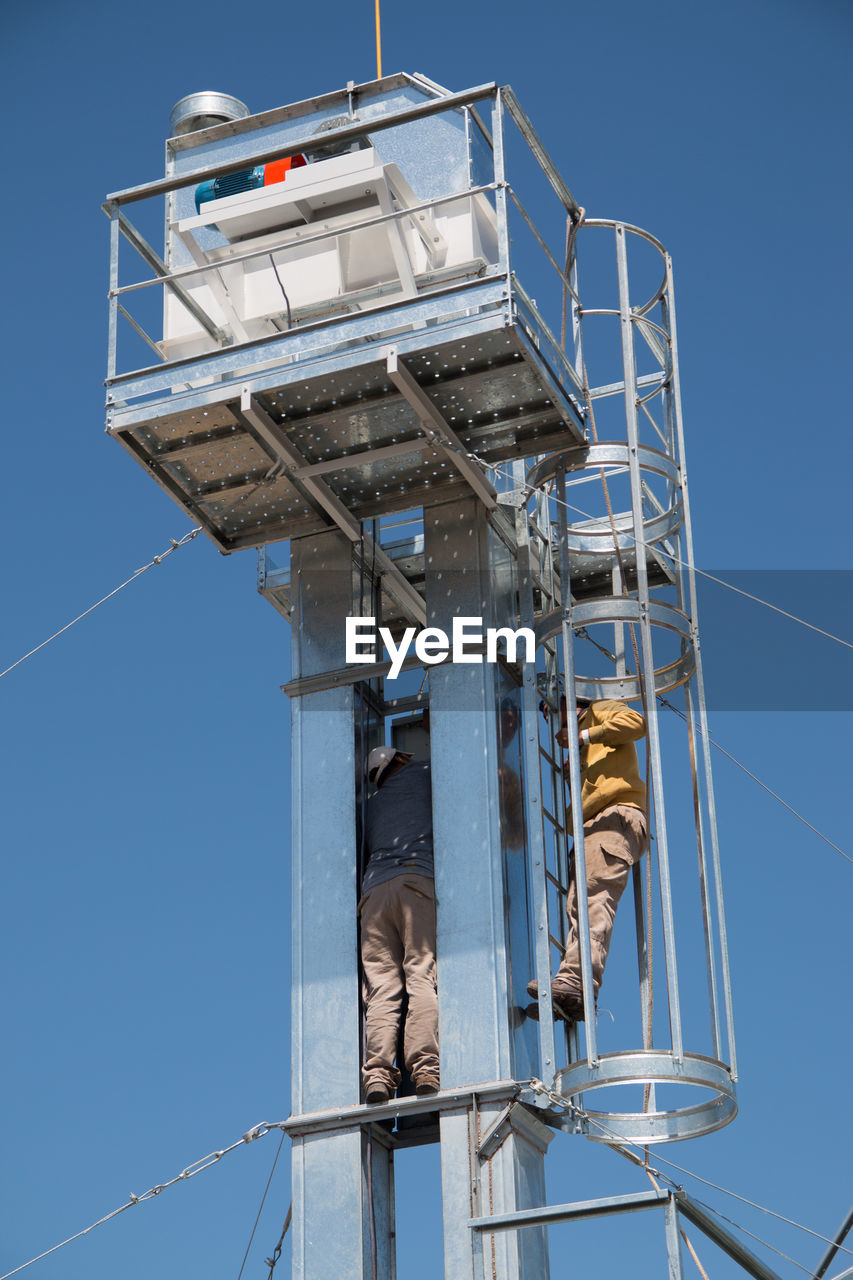 Low angle view of people working on tower against clear blue sky