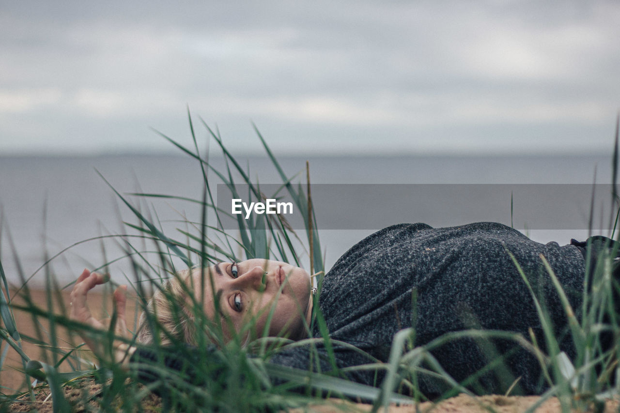 Portrait of young woman relaxing on sand at beach against sky