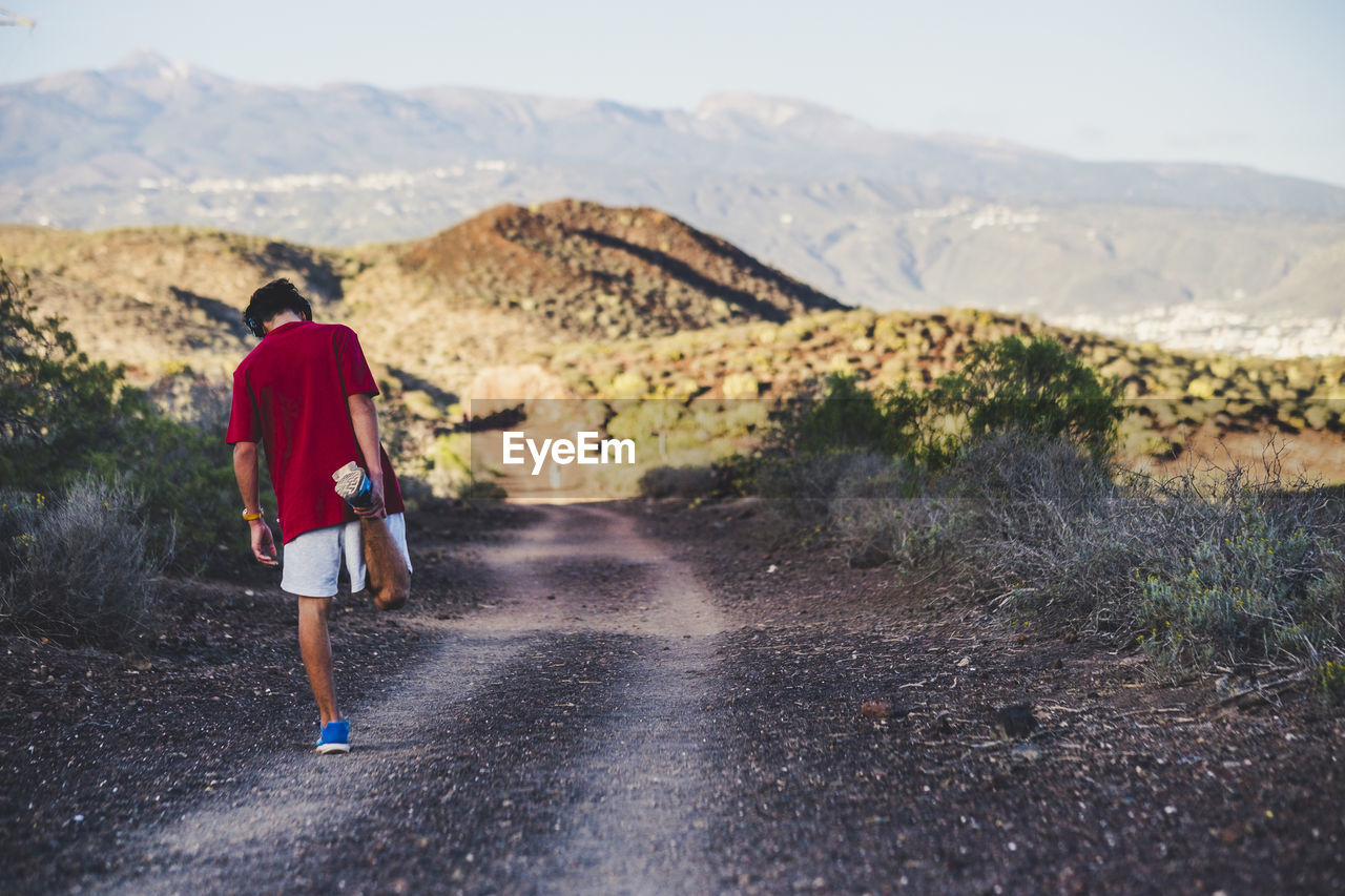 REAR VIEW OF MAN WALKING ON ROAD AGAINST MOUNTAINS