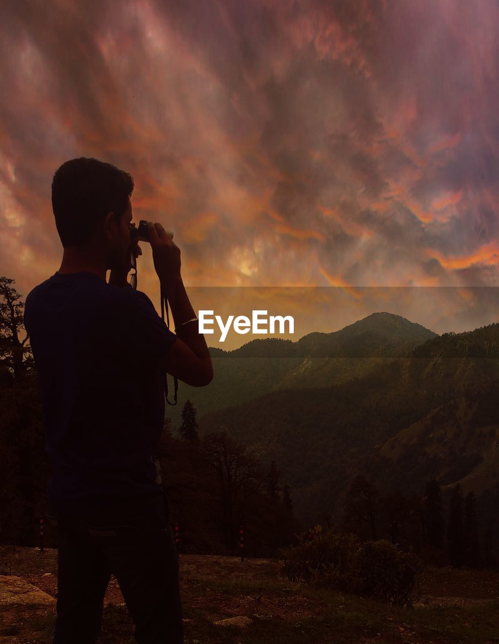 Rear view of man looking mountains through binoculars against cloudy sky during sunset