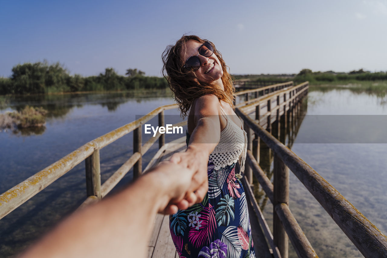 Woman holding hand of boyfriend while standing on footbridge against sky