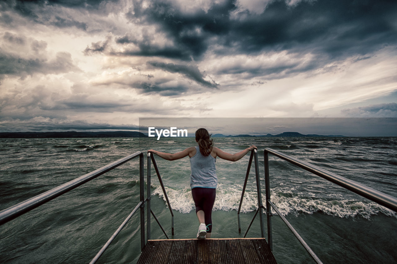 Rear view of woman moving down on pier in sea against cloudy sky