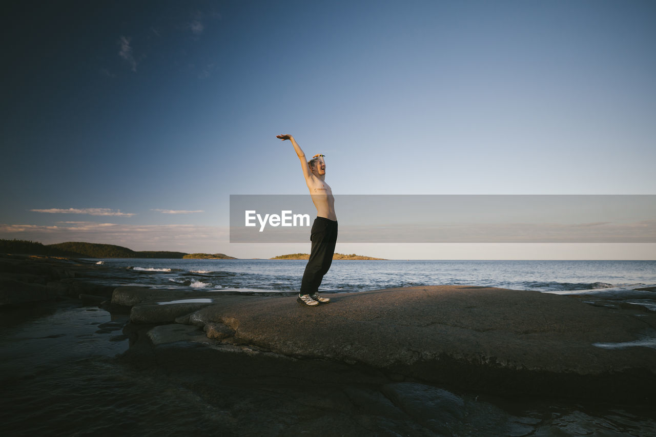 Shirtless transgender man standing on coast with raised arm