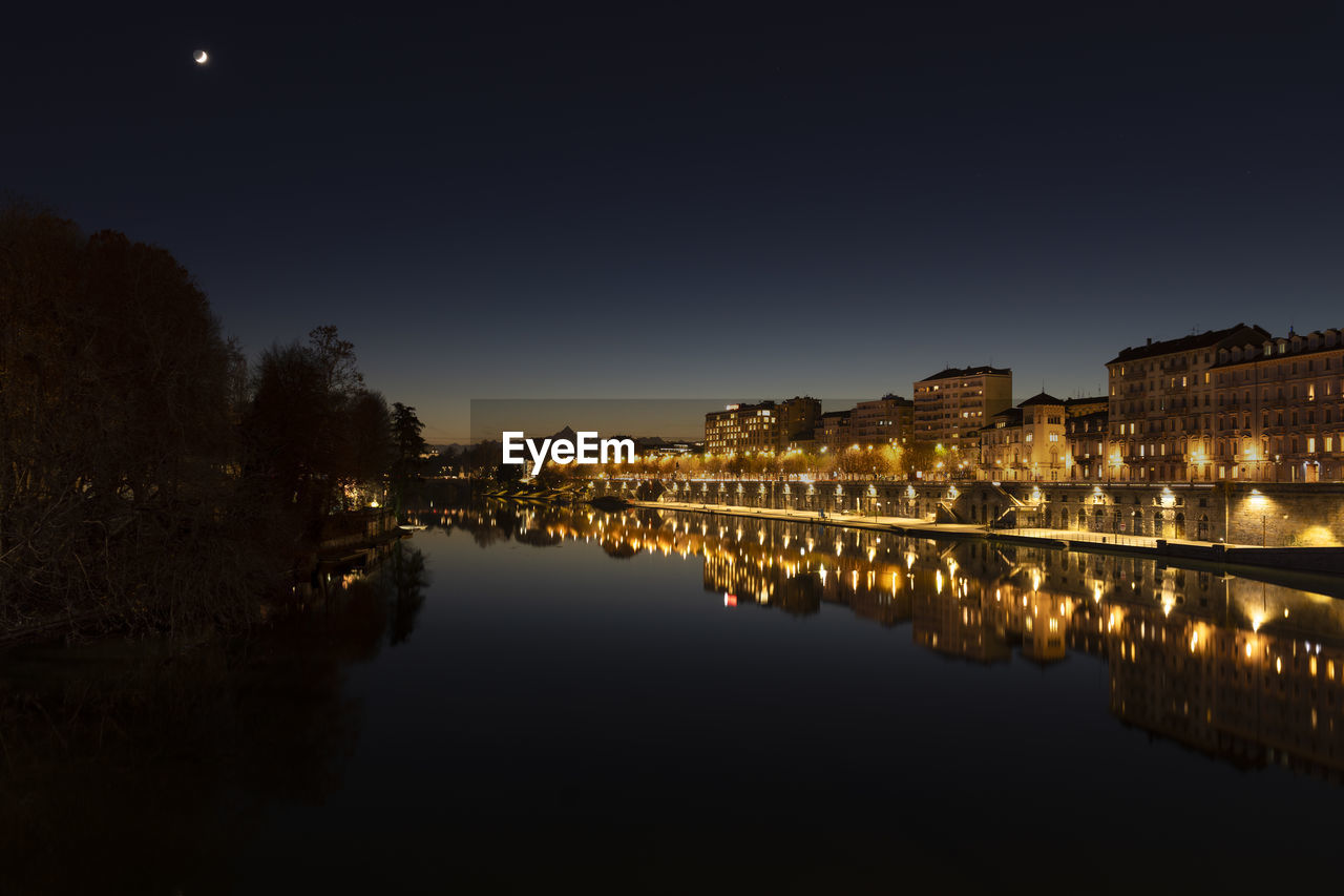 Illuminated buildings by lake against sky at night