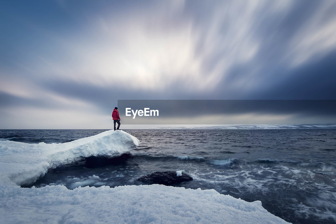 Man standing on snow covered rock by sea against cloudy sky