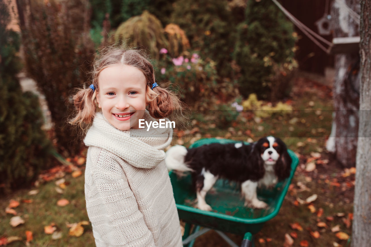 Portrait of smiling girl standing by dog in wheelbarrow on field