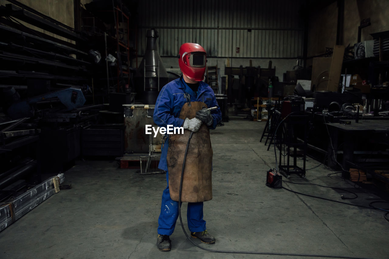 Anonymous mechanic in protective helmet and apron on blue overall standing with welding hose in light workshop near metal constructions person