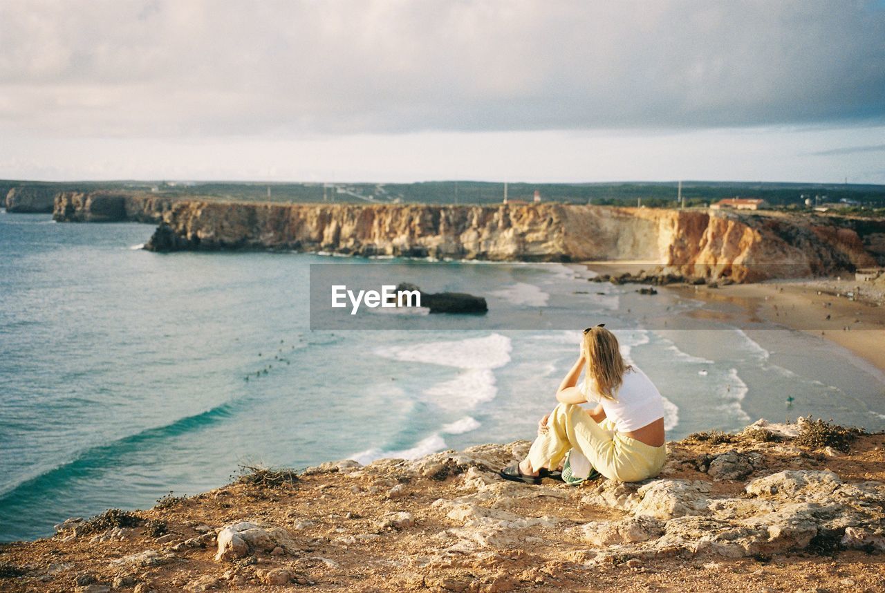 Rear view of woman sitting on rock by sea against sky