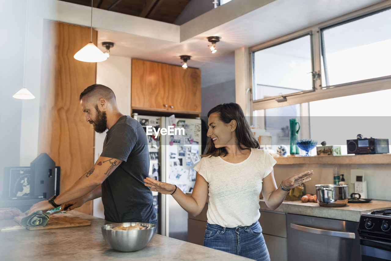 Happy woman gesturing while talking to man cutting vegetables in kitchen