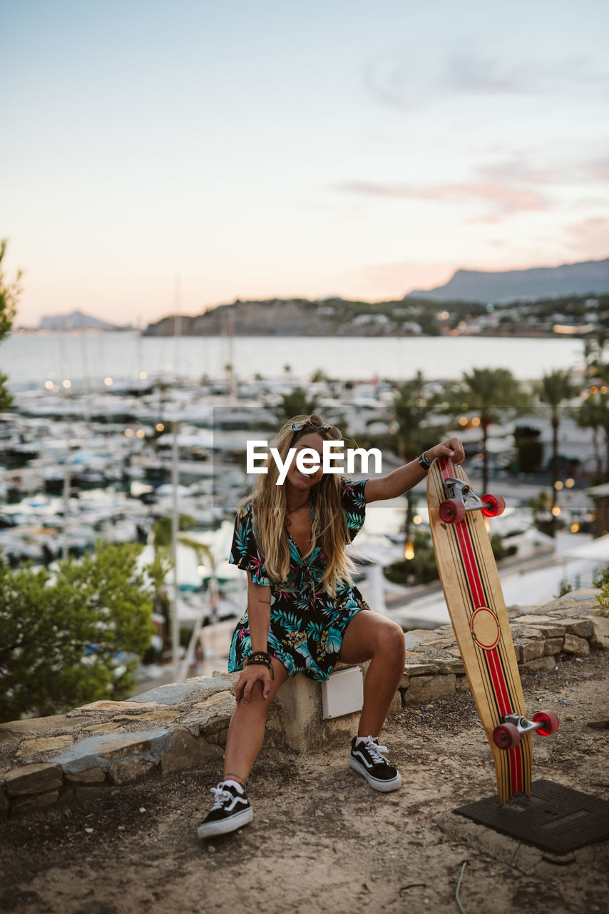 Young female with skate board in front of moraira's yacht club at dusk