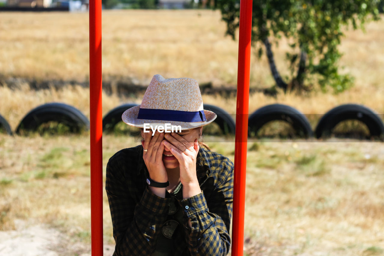 Young woman in checkered shirt and hat swinging on swing on playground. tired woman covering her 