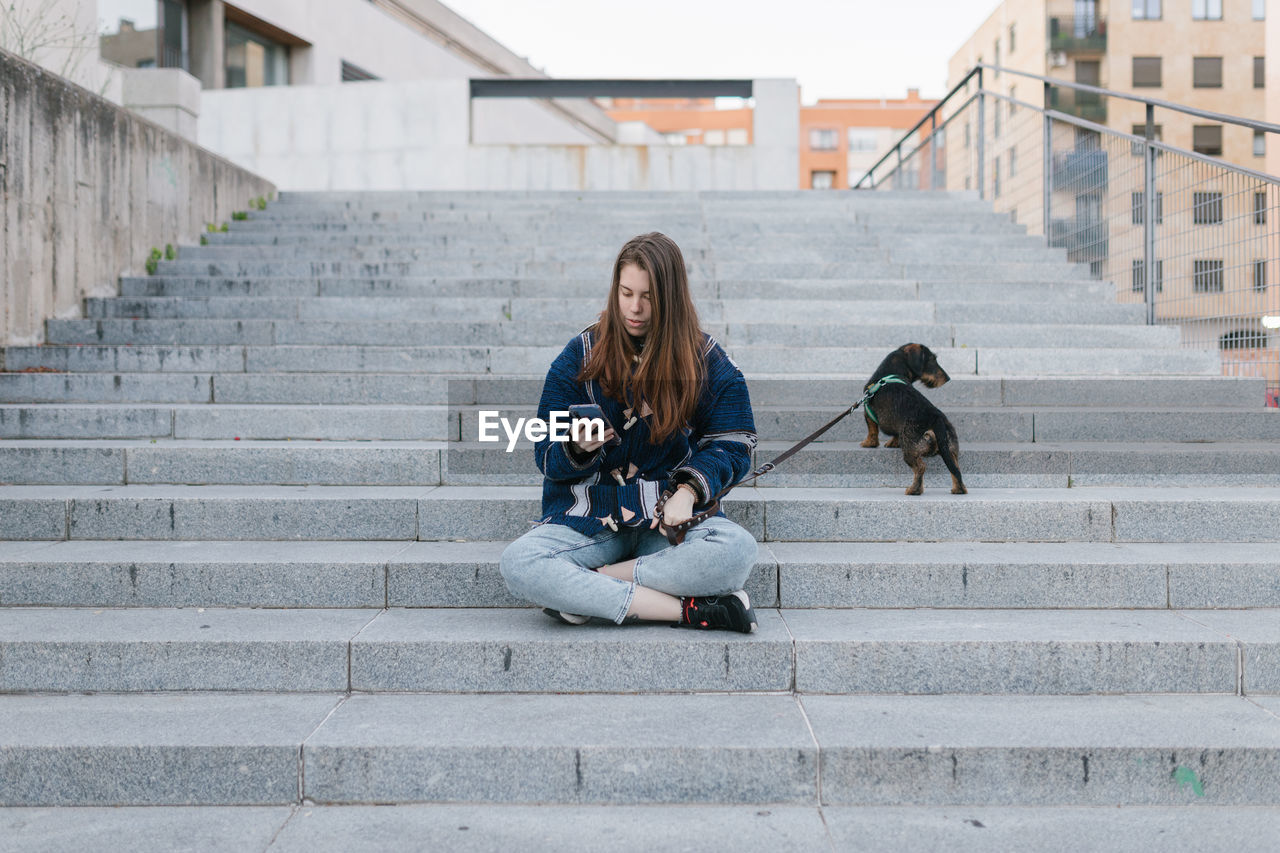 Young adult woman sit looking her phone and next to her dog after walk