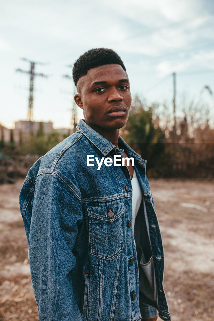 Young serious african american male with modern haircut in denim jacket looking at camera under cloudy sky in fall
