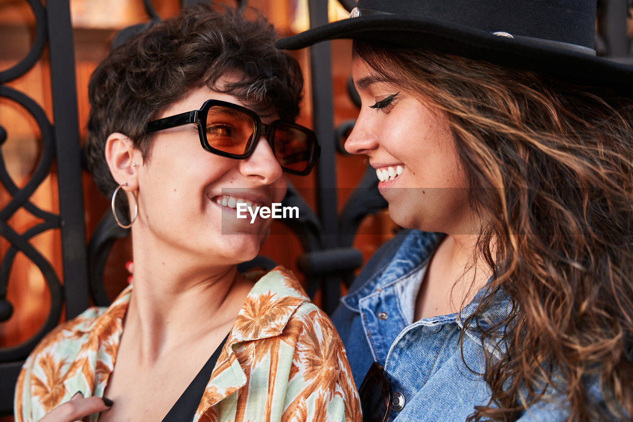 Happy young women in stylish clothes smiling and looking at each other against metal gate during romantic date on street of madrid, spain