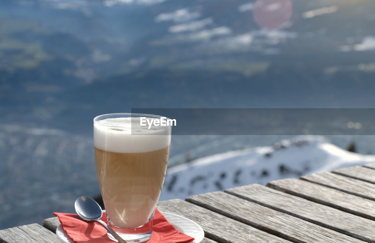 Glass of latte macchiato caffee latte on a wooden table and sunray. snow covered mountain background 