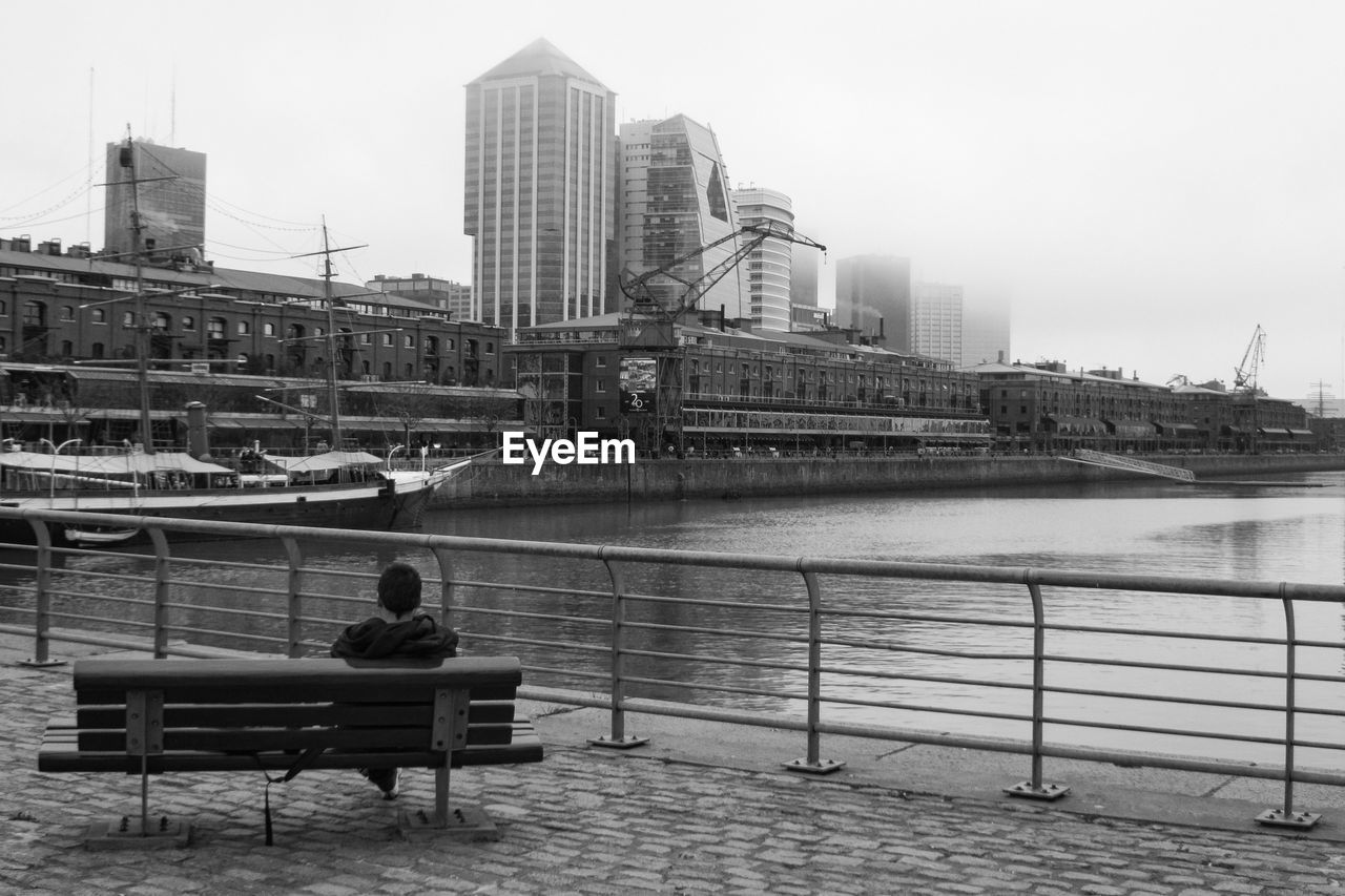 REAR VIEW OF MAN SITTING ON BENCH AT RIVERBANK