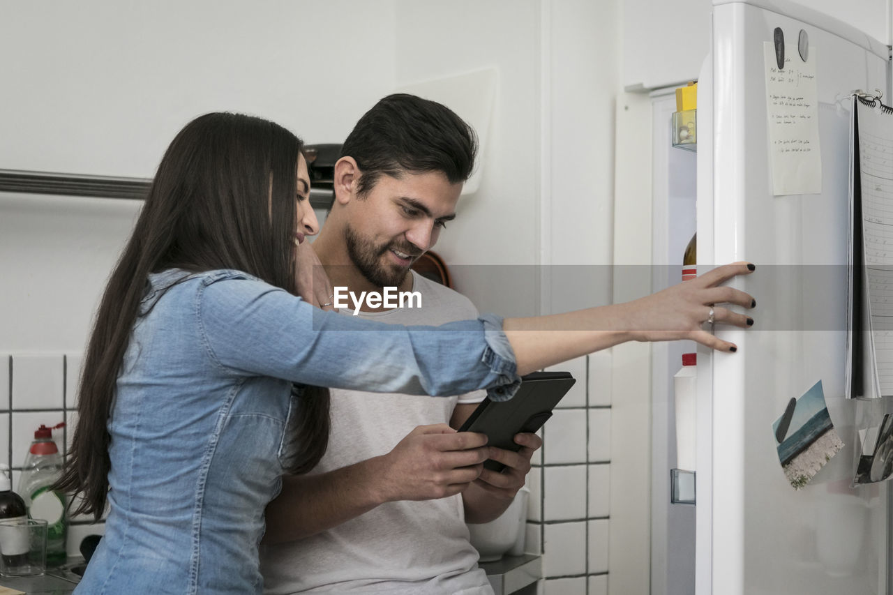 Smiling couple using digital tablet while standing by refrigerator at kitchen