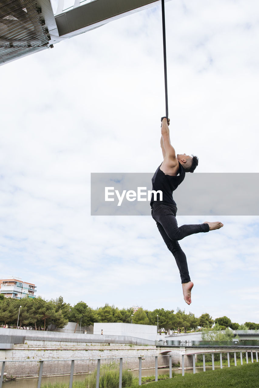 Low angle of fit male gymnast hanging on aerial straps during training in park