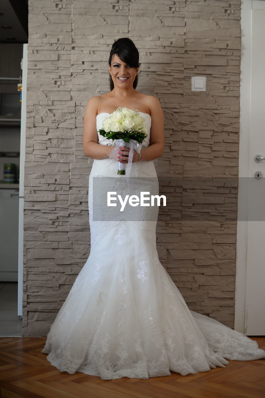 Full length of bride holding bouquet while standing against wall at home