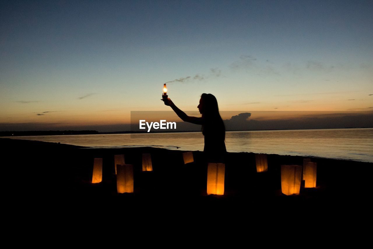 Silhouette woman holding illuminated electric lamp on beach at dusk