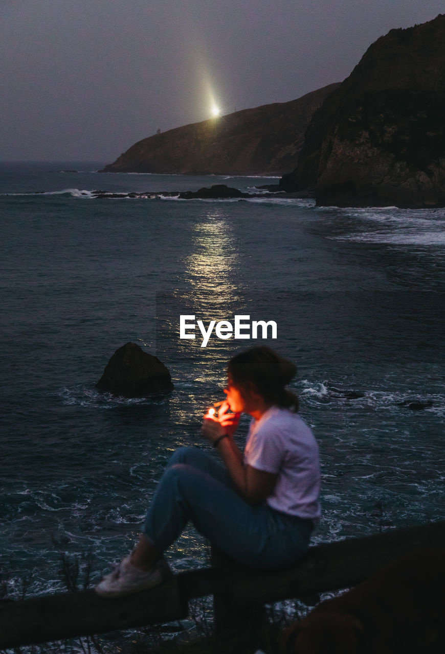 From above side view of casual pensive young female smoking cigarette while sitting on fence on rocky sea coast in summer evening in town of lekeitio in spain with beacon light in background