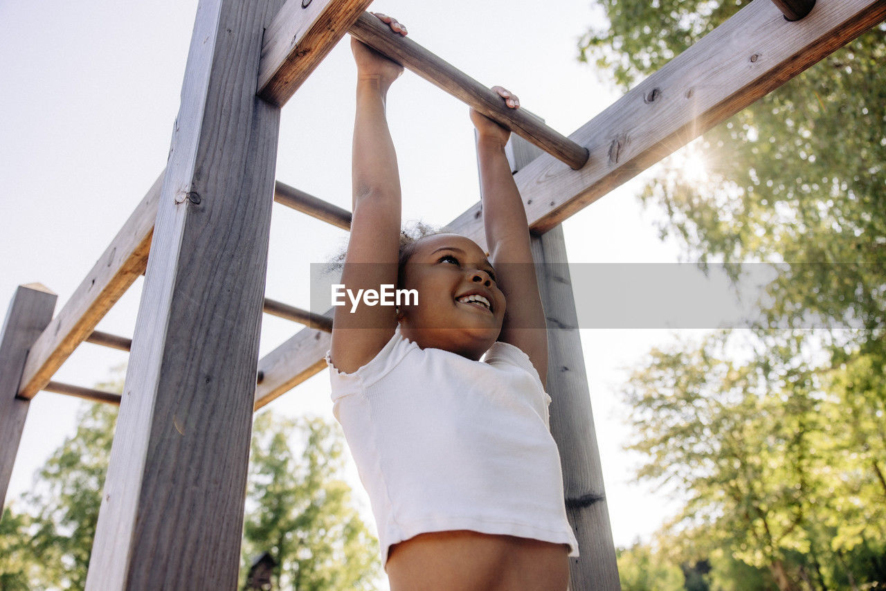 Low angle view of smiling girl hanging while doing monkey bars at summer camp