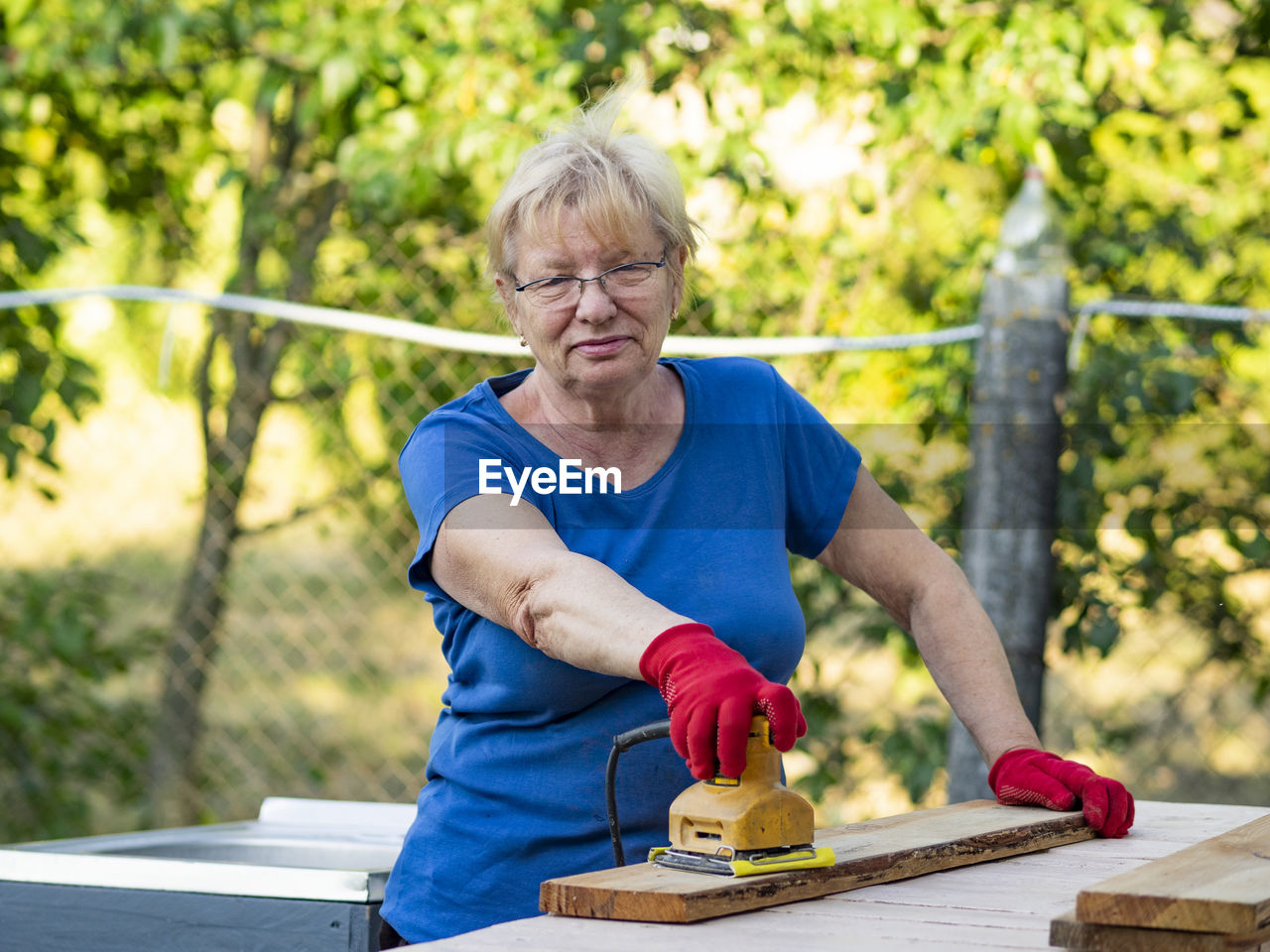 Senior caucasian woman in a blue t-shirt and red gloves is sanding a board with a sander 