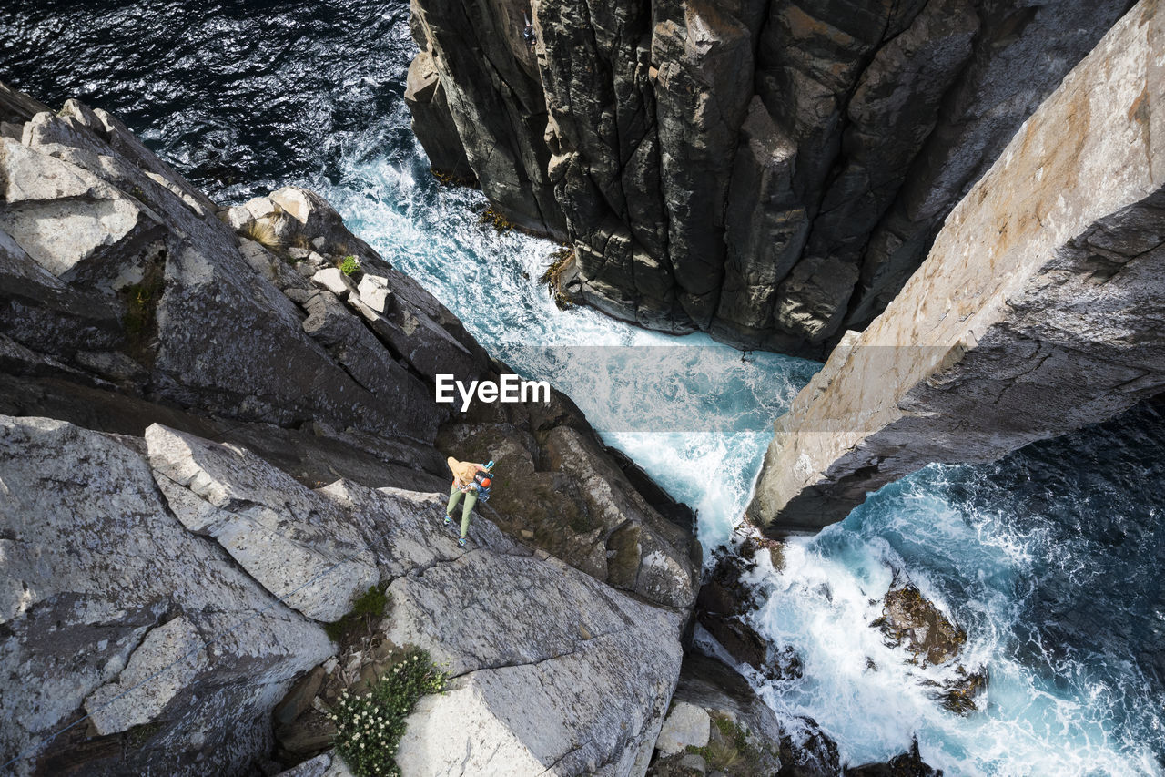 Female rockclimber looks down towards the crashing waves as she rappels down from the edge of a sea cliff to the bottom of the totem pole, a rock column which emerges out of the ocean