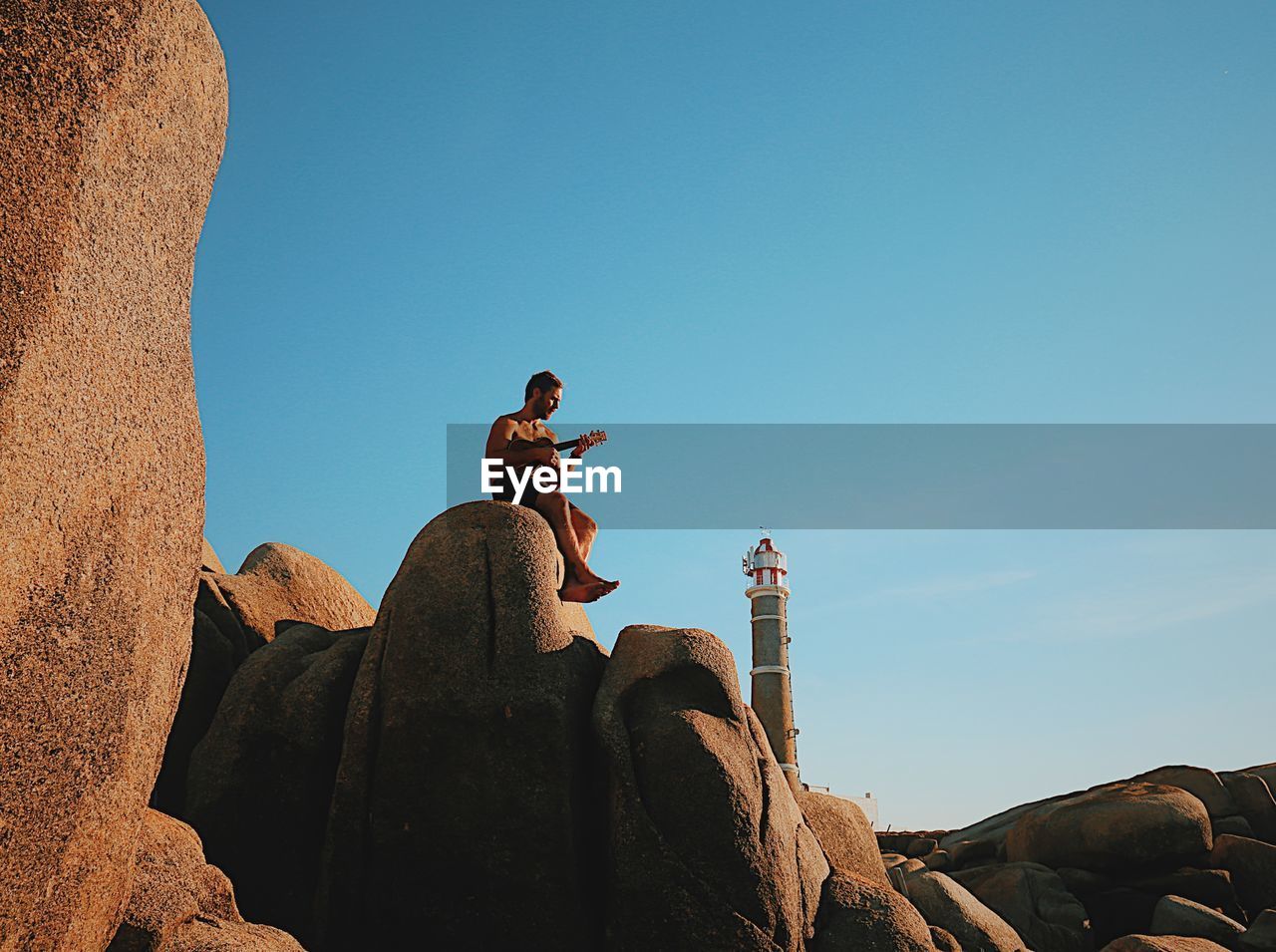 Low angle view of mid adult man playing guitar while sitting on rock formation against blue sky