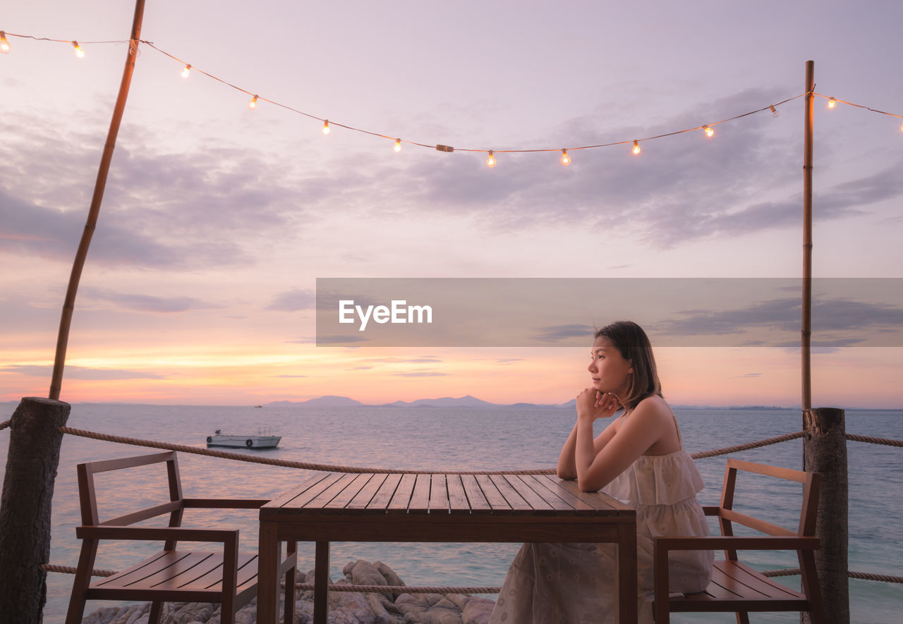 Side view of woman sitting by sea on bench during sunset