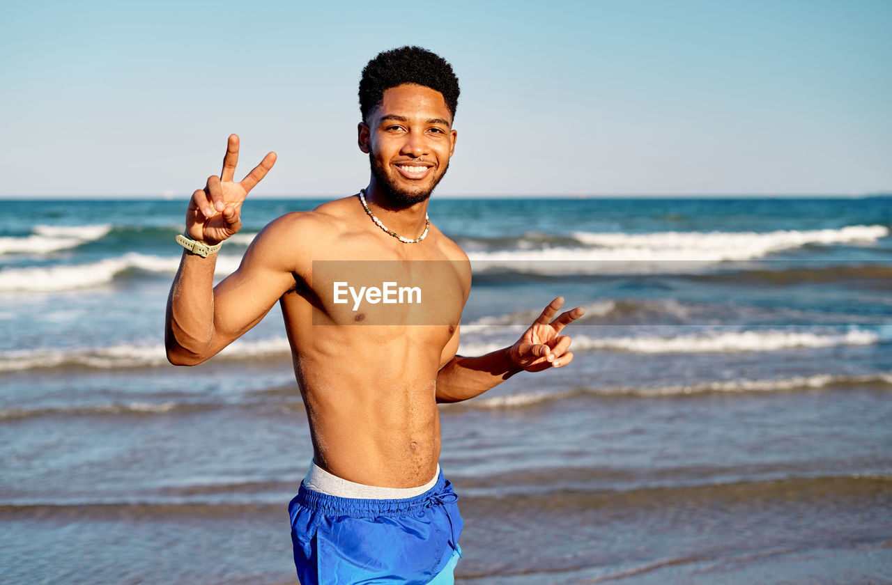Glad shirtless black male tourist looking at camera with smile and gesturing v sign against waving sea and cloudless sky on weekend day on beach