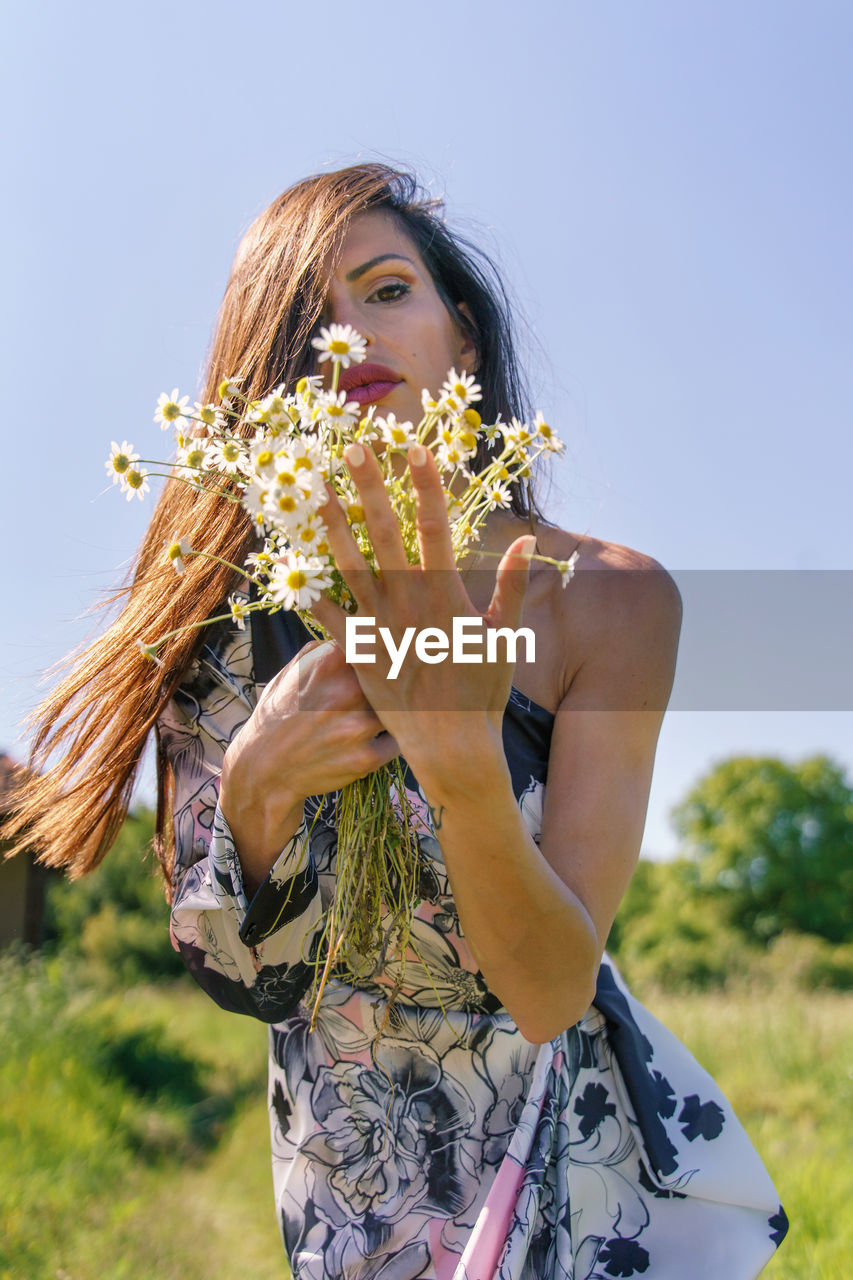 MIDSECTION OF WOMAN HOLDING FLOWERING PLANT IN FIELD