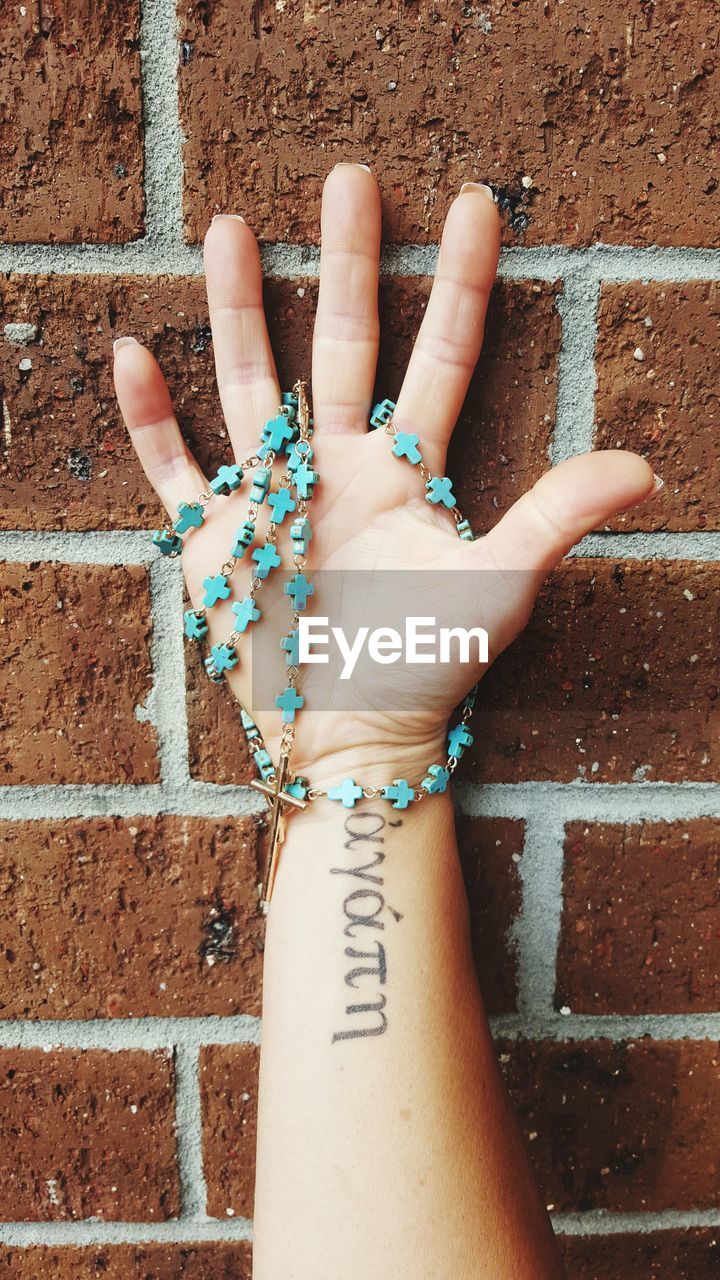 Cropped image of tattooed hand holding rosary beads against brick wall
