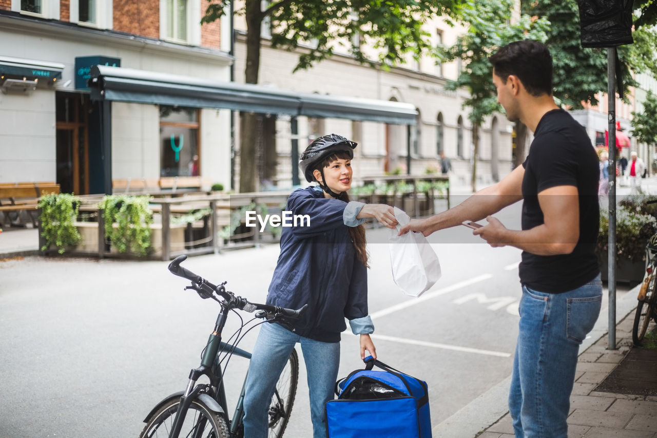 Food delivery woman giving package to male customer standing on sidewalk in city