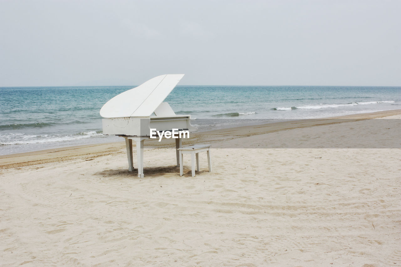 White piano and stool on sandy beach against clear sky